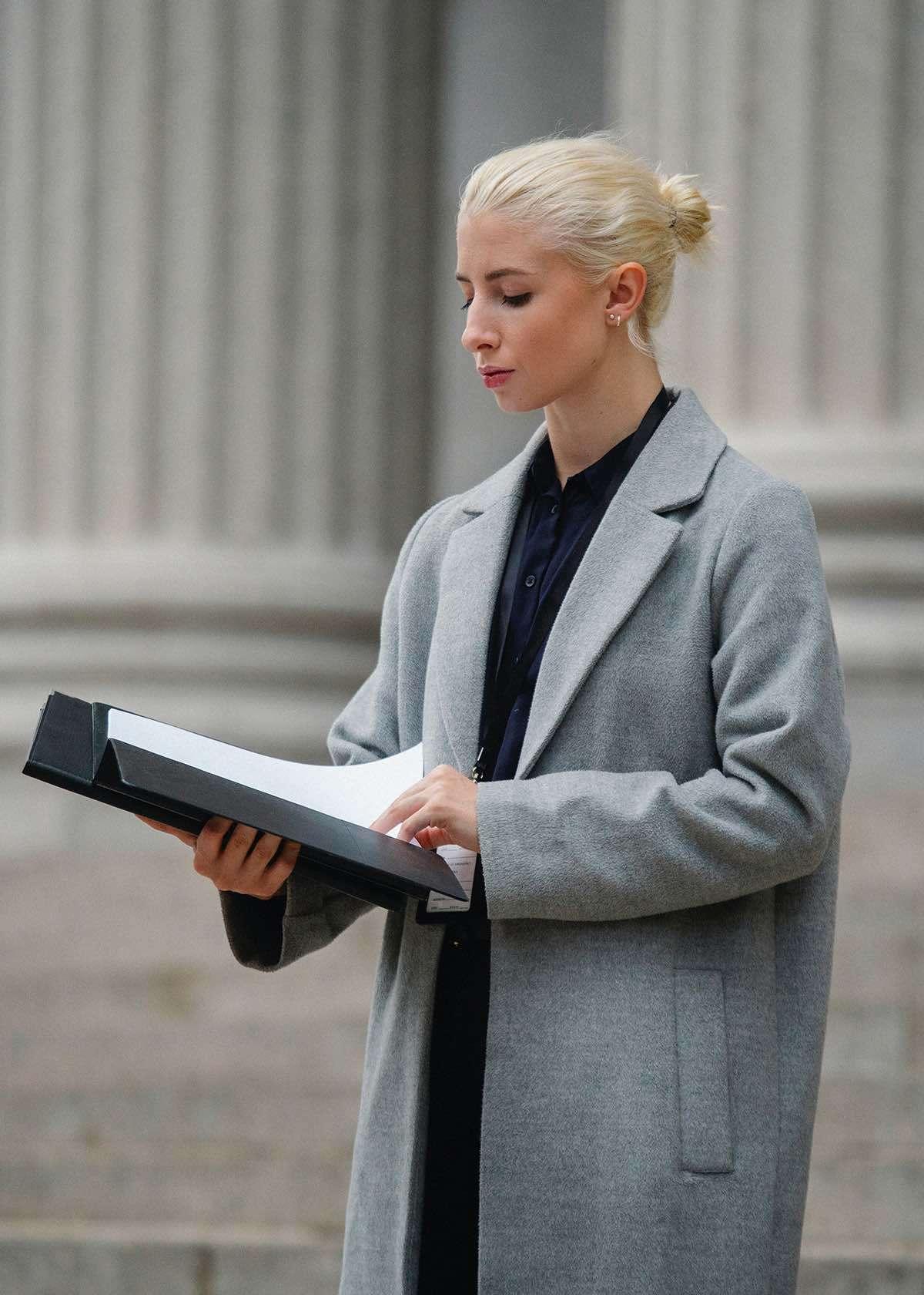 Investigatore privato donna con capelli biondi, indossa una camicia nera e un cappotto grigio legge delle documentazioni