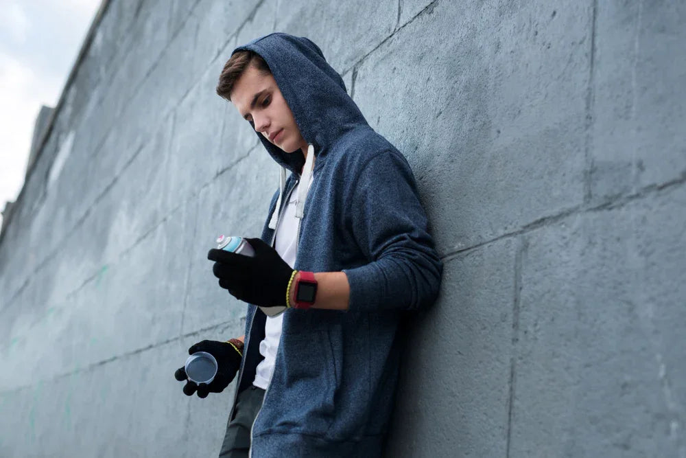 Giovane ragazzo in felpa con cappuccio e guanti tiene in mano una bomboletta spray con l' intenzione di imbrattare il muro su cui è appoggiato, necessita di indagini di controllo minori per attività illecite o comportamenti criminali.
