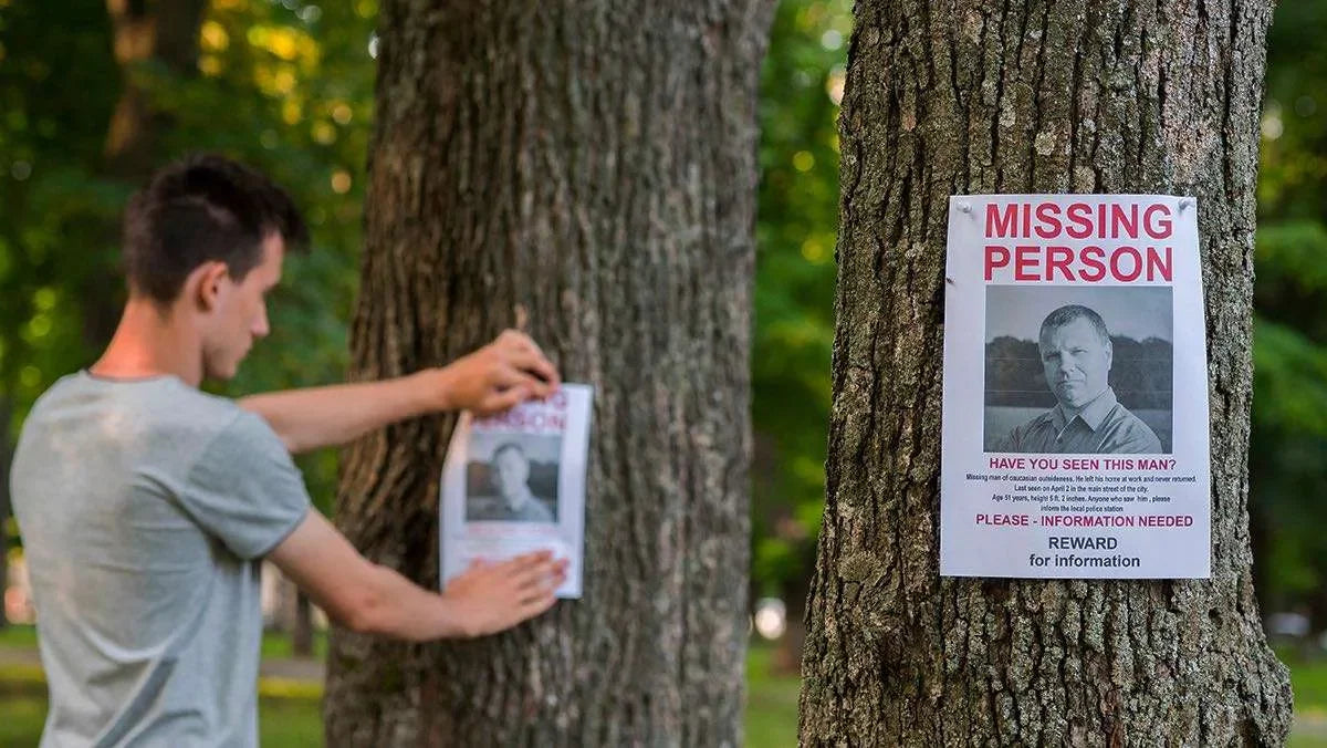 Ragazzo affissa sul tronco di due alberi un poster contenente la foto di un' uomo e il testo di annuncio persona scomparsa. Simbolo di Indagini e ricerche persone scomparse.