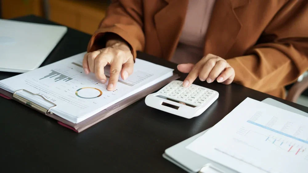 Donna seduta dietro ad una scrivania compie un' analisi finanziaria con documenti e calcolatrice per verifica patrimoniale preventiva e indagini economiche.