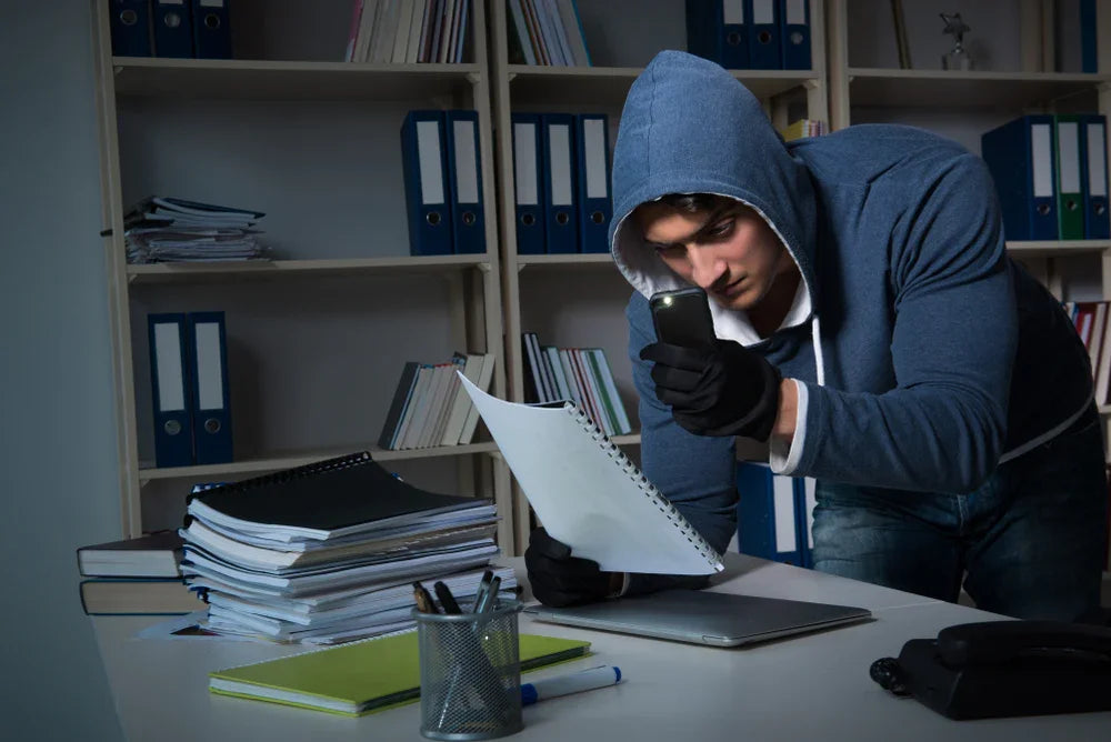 Uomo con felpa e guanti fotografa documenti aziendali riservati, simbolo di furto e accessi sospetti.