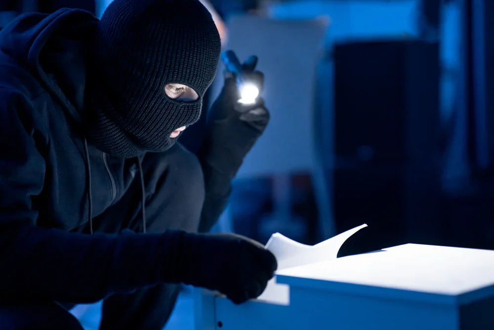 Ladro vestito totalmente di nero con passamontagna e torcia in mano apre un cassetto e legge un documento. Simbolo di indagini per violazione di brevetti o segreti industriali.