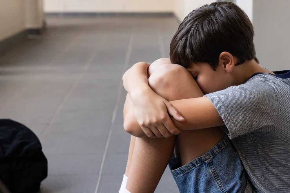 Ragazzino seduto e accovacciato a terra da solo con la testa tra le ginocchia, simbolo di bullismo scolastico invisibile ma costante.