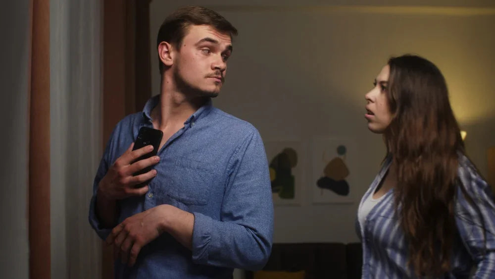 Uomo nasconde il proprio telefono appoggiandolo sul proprio petto alla propria partner che lo guarda con espressione sospetta. Simbolo di Frequentazioni o comportamenti incoerenti