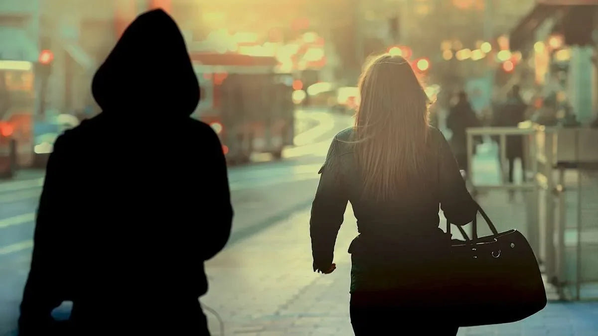 In ambiente esterno urbano, una ragazza di spalle con borsa viene seguita da persona non identificata incappucciata e vestita di nero.
Simbolo di indagini di controllo anti-stalking.