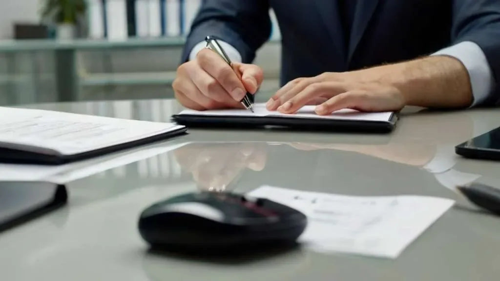 Mani di un uomo in giacca che scrive su un foglio appoggiato ad una scrivania apparecchiata di mouse, telefono e documenti. Simbolo di indagini per controllo minori.