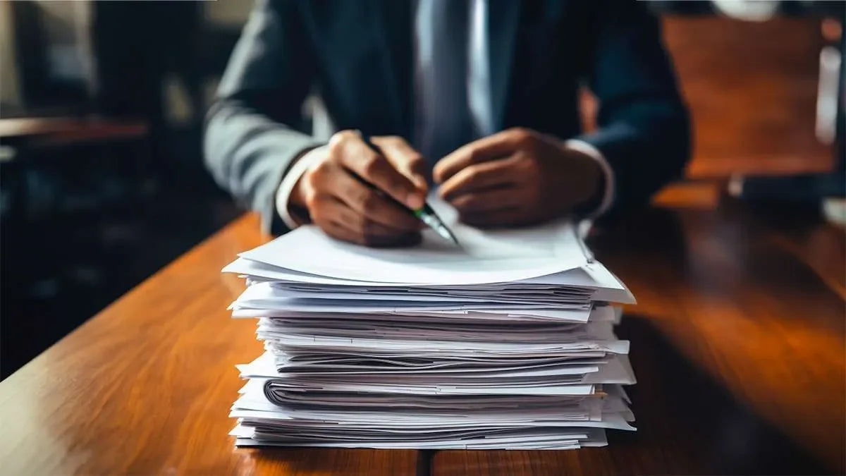 Mani di un uomo in giacca e cravatta che stringono una penna appoggiate ad una pila di fogli e documenti. Sinonimo di indagini patrimoniali.