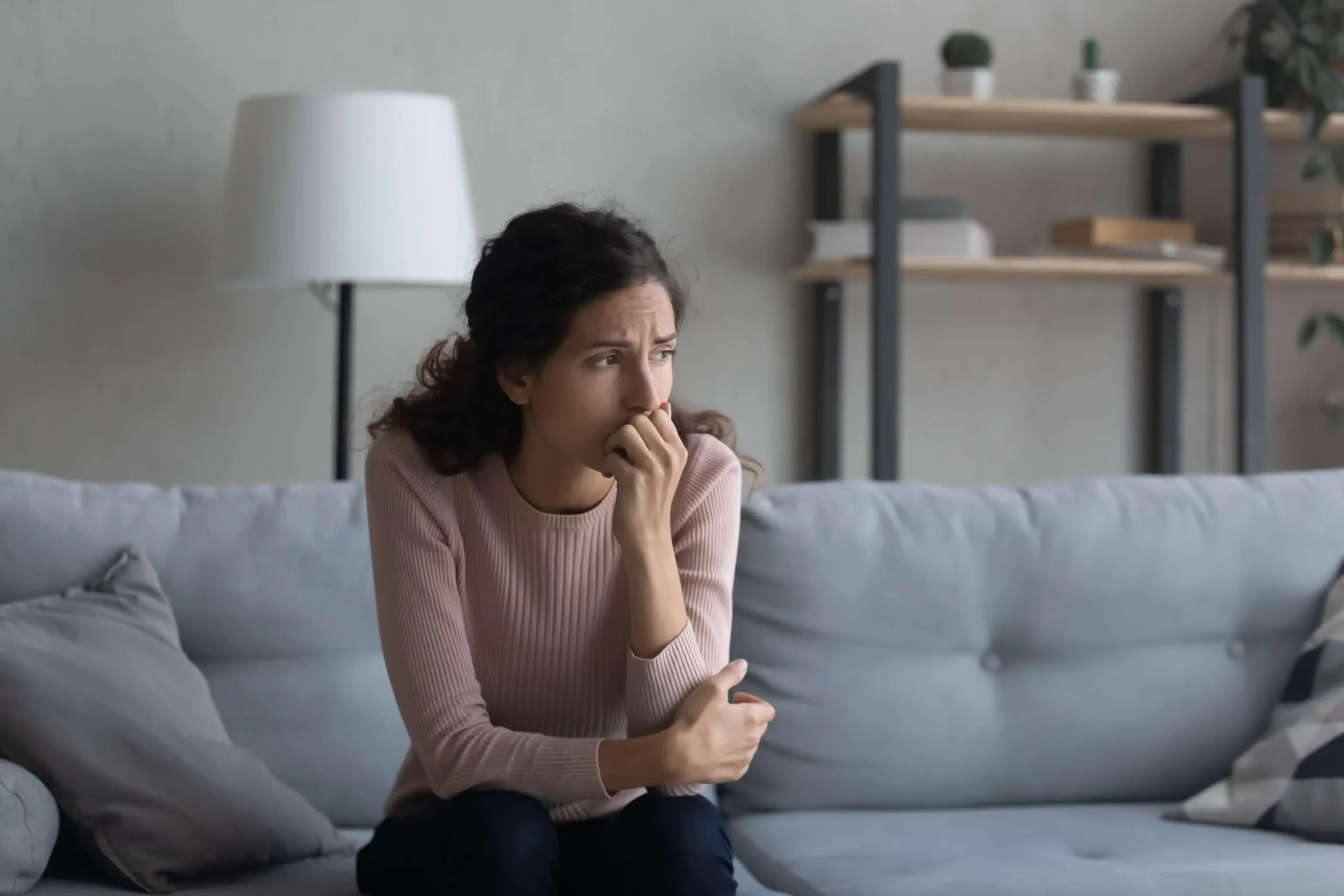Donna in maglia rosa seduta sul divano con mano davanti alla bocca seduta sul divano di casa con espressione preoccupata. Simbolo di indagini di infedeltà coniugale.