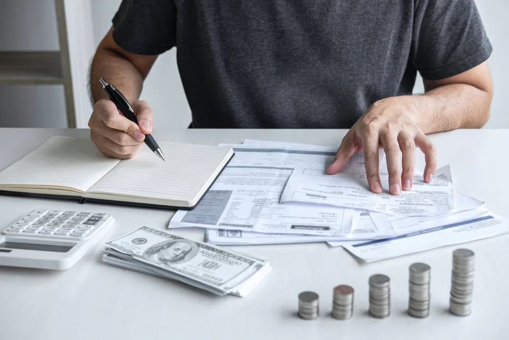 Uomo con penna in mano seduto ad una scrivania apparecchiata di agenda, documenti, monete e contanti. Simbolo di indagini di verifica patrimoniale.